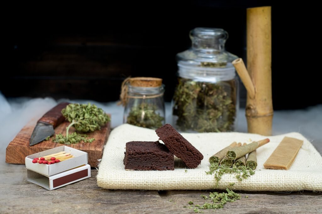 Pre-Roll marijuana joints and cake brownies with curing cannabis buds in a clear glass jar laying