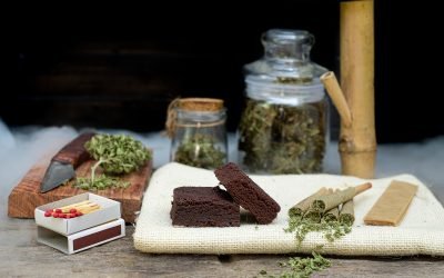 Pre-Roll marijuana joints and cake brownies with curing cannabis buds in a clear glass jar laying